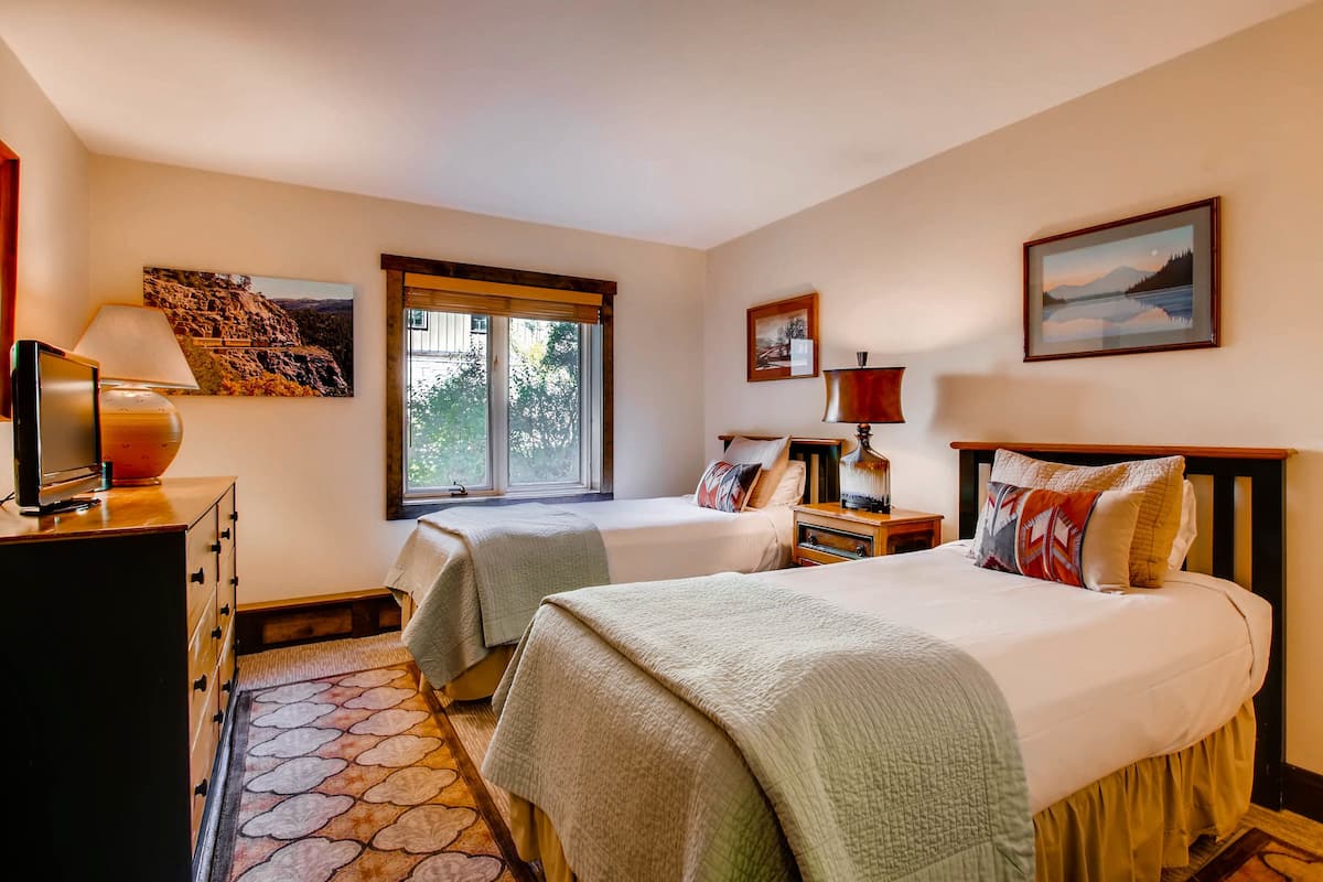 A bedroom with two beds, a dresser, a TV, and a window with a view of trees.