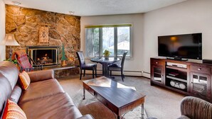 A living room with a stone wall, fireplace, leather sofa, coffee table, TV, and a stone fireplace mantel. - Westwind at Vail (Vail)