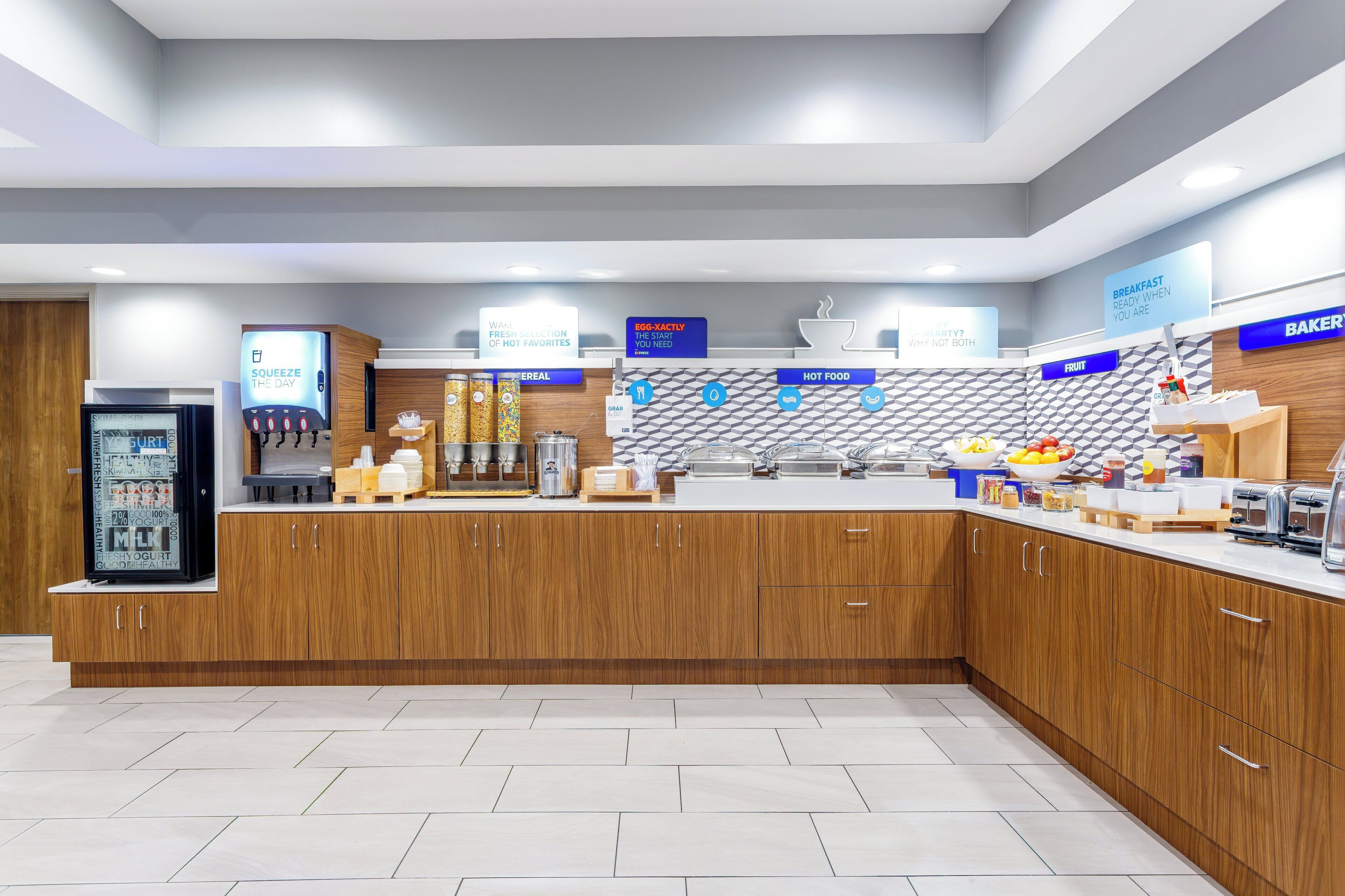 A modern breakfast bar with wooden cabinets and a white countertop. There are signs indicating breakfast options and a refrigerator with yogurt.
