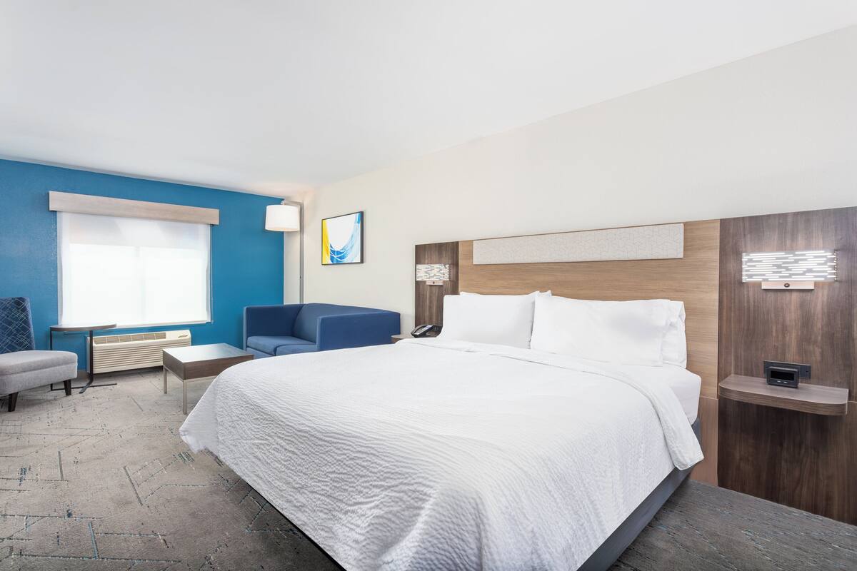 Una habitación de hotel con una cama grande, un escritorio, una silla, una mesita, una mesita de noche, una lámpara y una ventana.
