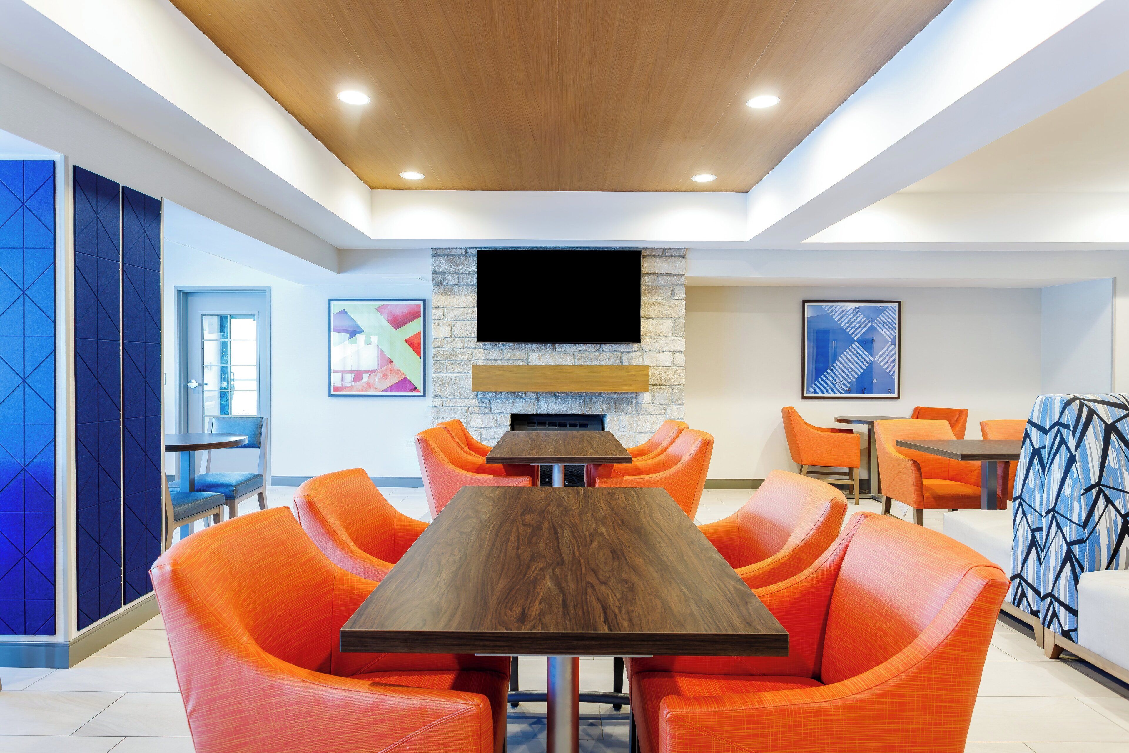 A modern dining area with a central wooden table surrounded by orange chairs, a stone fireplace, and a flat-screen TV mounted above.