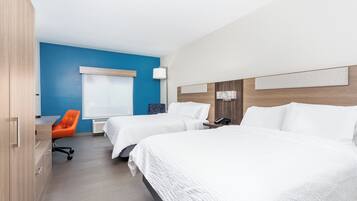 A hotel room with two beds, a desk, an orange chair, a window with blinds, and a lamp.