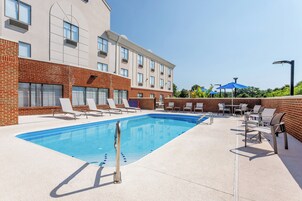Piscina com espreguiçadeiras e guarda-sóis, parede de tijolos e um edifício com várias janelas.