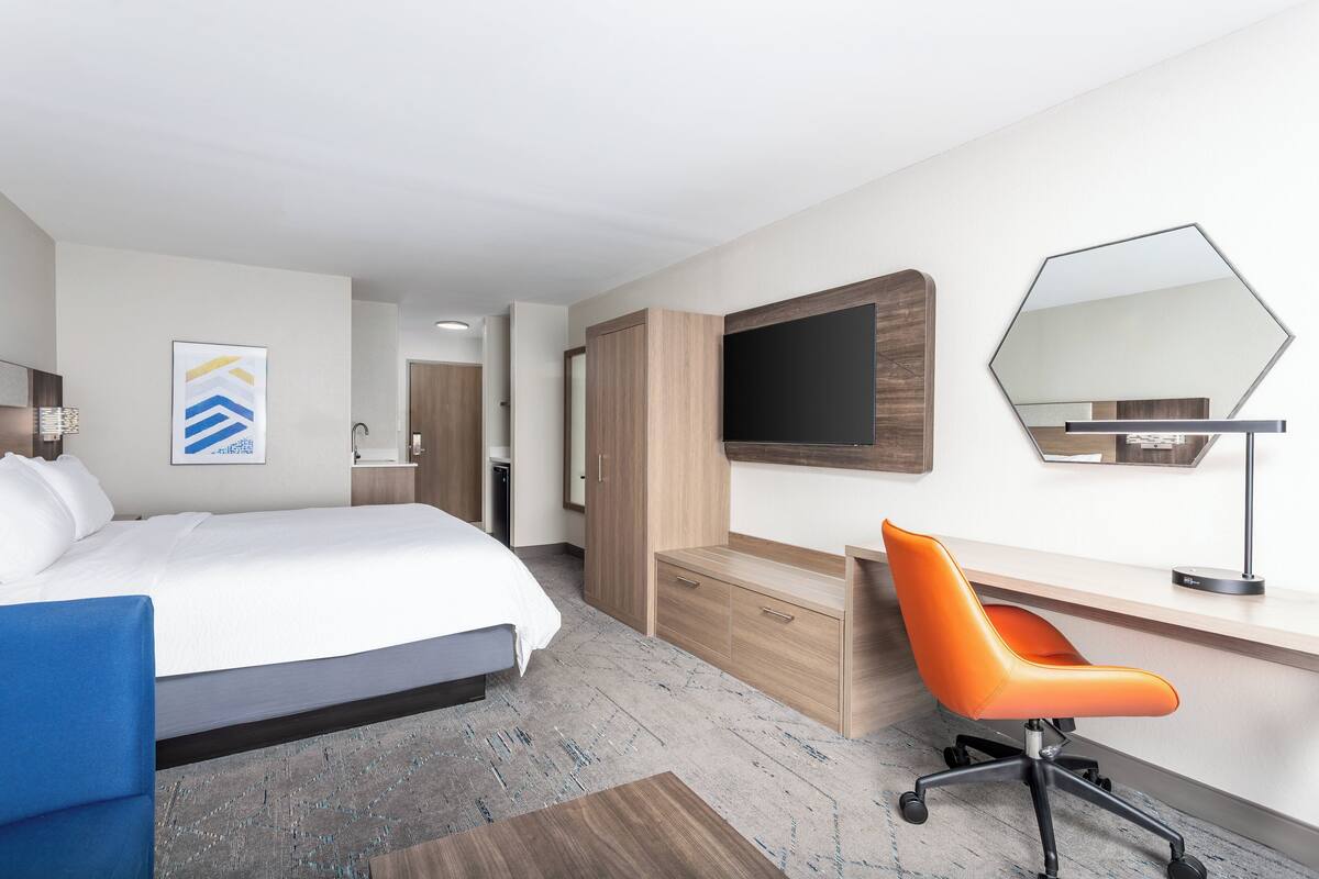 Habitación de hotel con cama, escritorio, una silla naranja, un espejo grande y televisor.