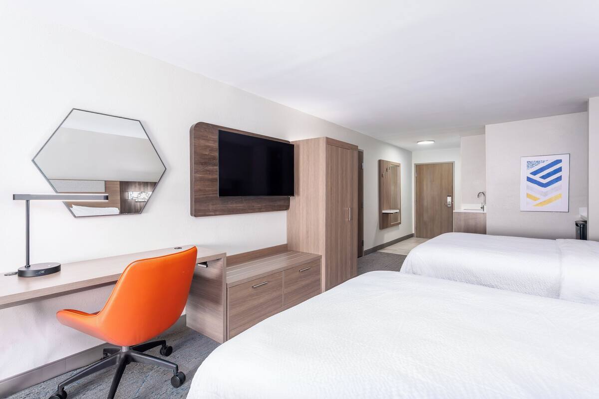 Una habitación de hotel con escritorio, una silla de oficina naranja, televisión, armario y dos camas.