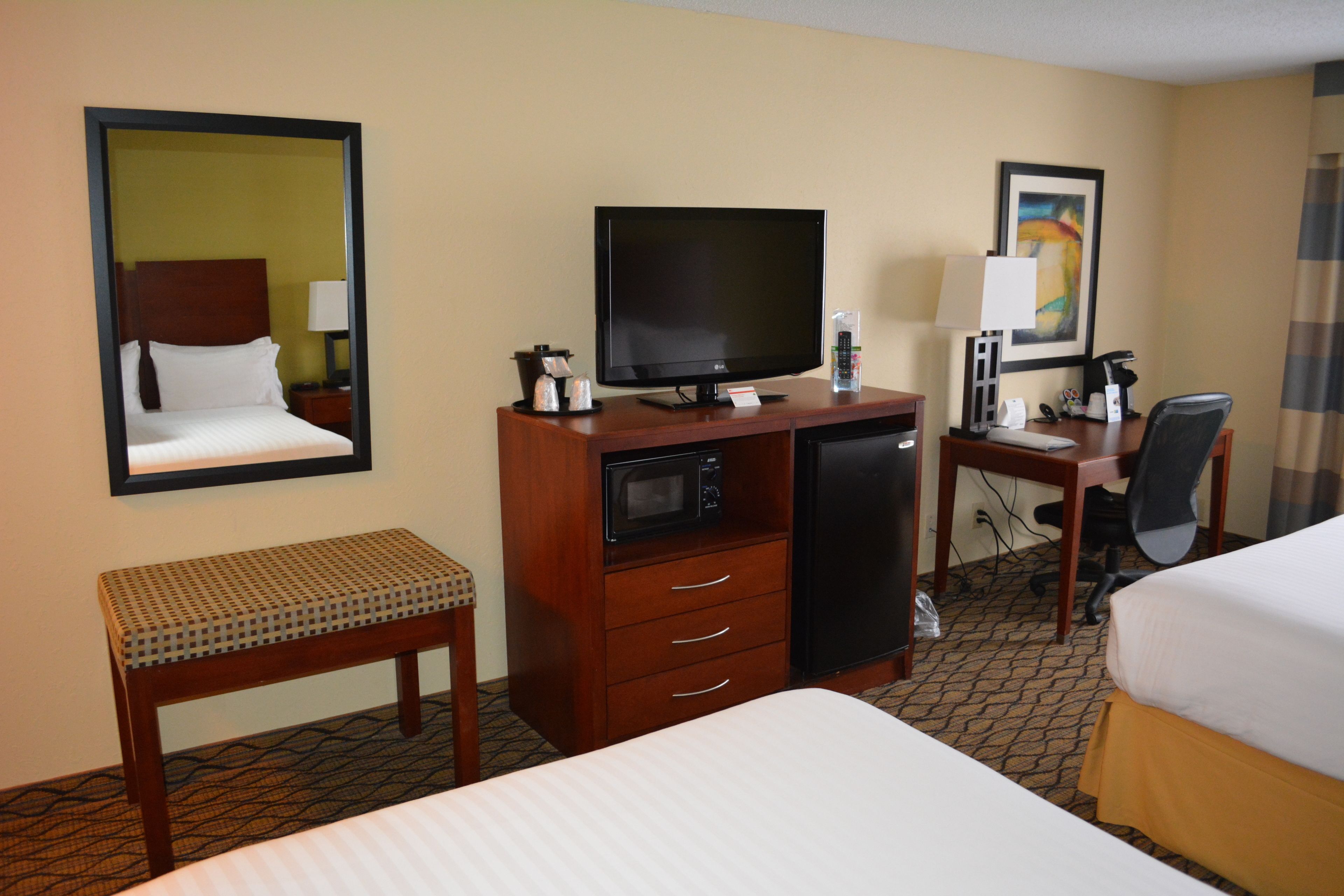 A hotel room with a bed, a desk with a computer, a television, a microwave, and a small bench.