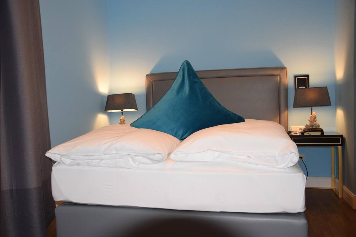 Un lit bien fait, avec du linge de lit blanc et un coussin turquoise, deux lampes de chevet et une petite table avec une lampe.