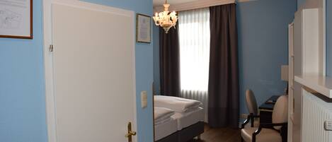 Un dormitorio con cama, un escritorio y una silla. Se observa una araña, una puerta y cortinas.