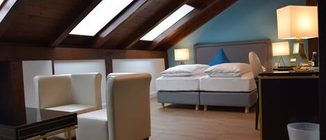 Una habitación de hotel con techo inclinado, cama con almohadas, mesita de noche, lámpara y una silla.
