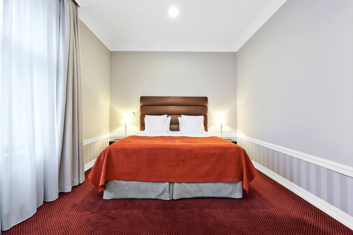 Una cama bien hecha con una colcha roja y almohadas blancas, un cabecero marrón y un suelo cubierto de alfombra roja.