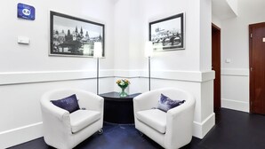 En moderne hotellobby med to hvide lænestole, et lille bord med en vase, og to sort-hvide fotografiudstillinger af bybilleder på væggene.