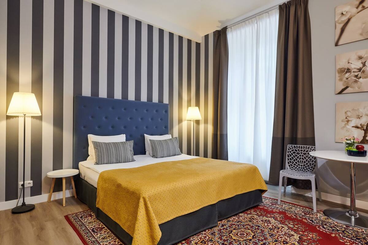 Una habitación de hotel con pared a rayas, cabecera acolchada, cama con manta amarilla, dos lámparas de noche, una mesita redonda con una silla y un tapete estampado.