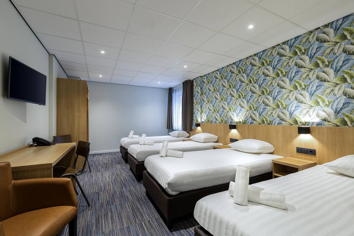 A hotel room with four single beds, a wooden desk, a flat-screen TV, and a large floral-patterned wall.