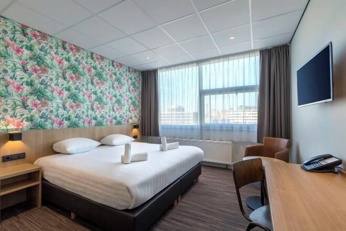 A hotel room with a large bed, a desk, a chair, a TV, and a window with a view of the city.