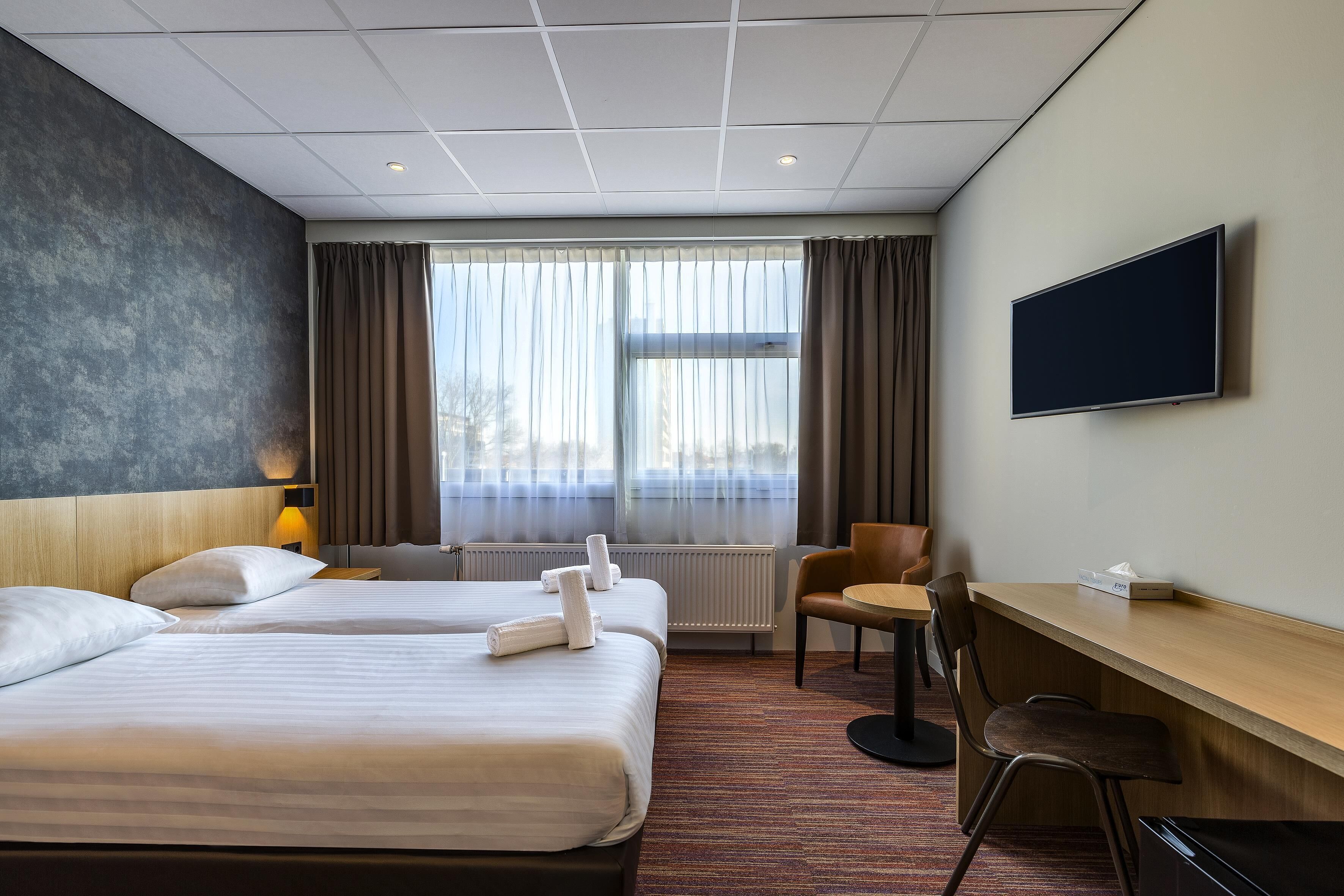 A hotel room with two beds, a desk, a chair, a TV, and a window with curtains.