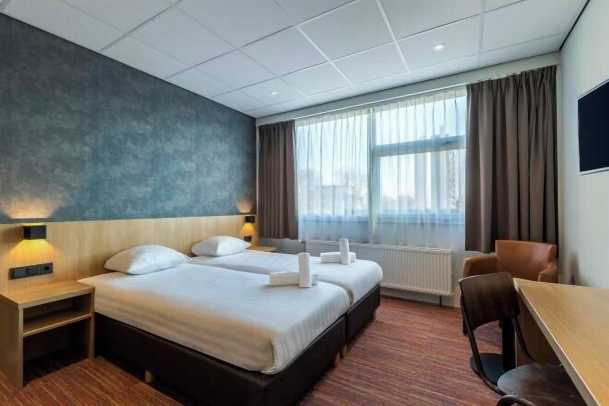 A hotel room with two beds, a desk, a chair, a window with curtains, and a radiator.