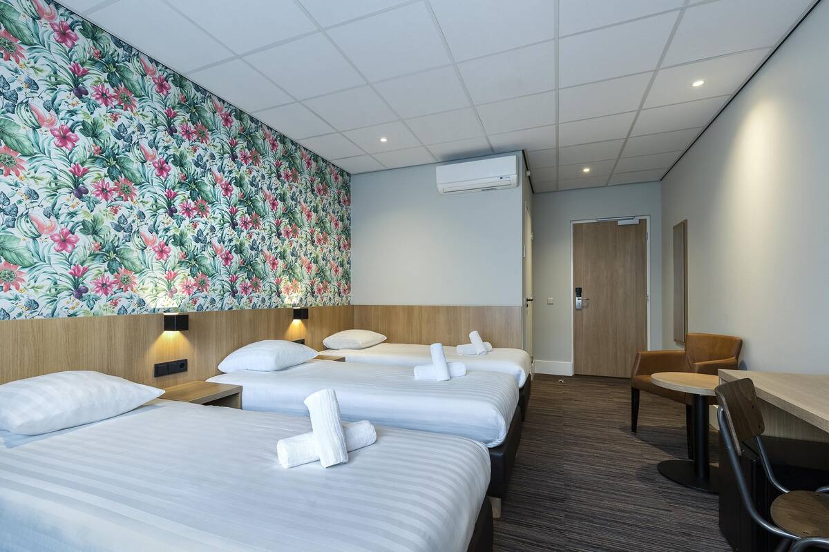 A hotel room with three beds, a wooden headboard, a floral wallpaper, a desk with a chair, and an air conditioning unit.