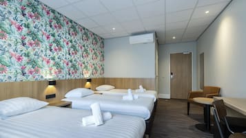 A hotel room with three beds, a wooden headboard, a floral wallpaper, a desk with a chair, and an air conditioning unit.