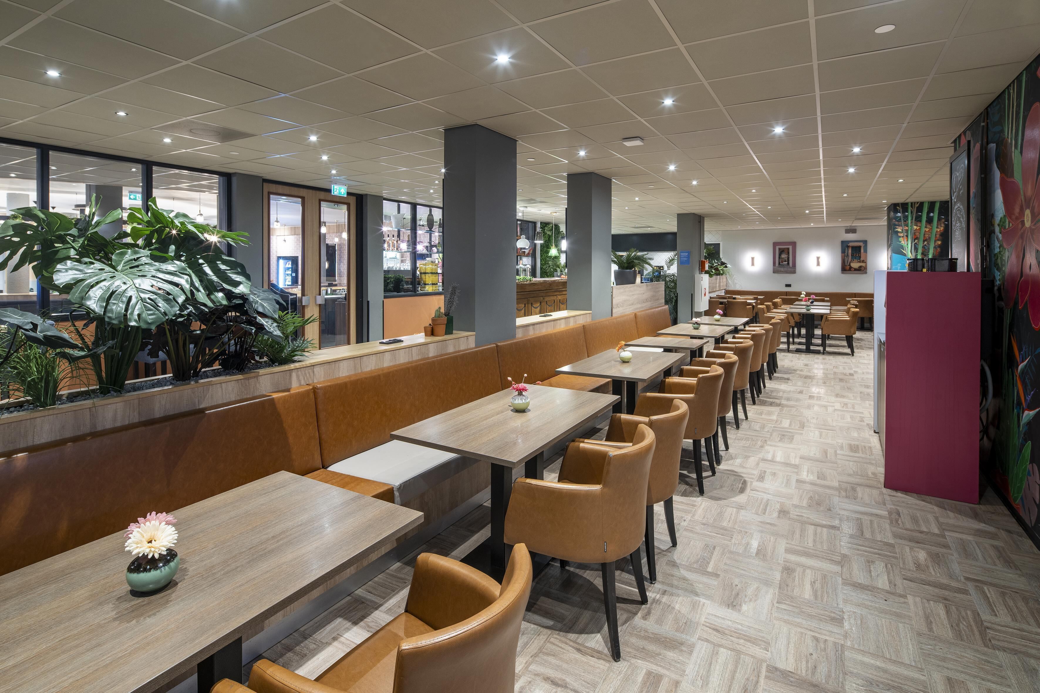 A modern restaurant with long wooden tables and cushioned booths, a tiled floor, and large windows offering views of the outside.