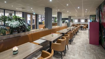 A modern restaurant with long wooden tables and cushioned booths, a tiled floor, and large windows offering views of the outside.