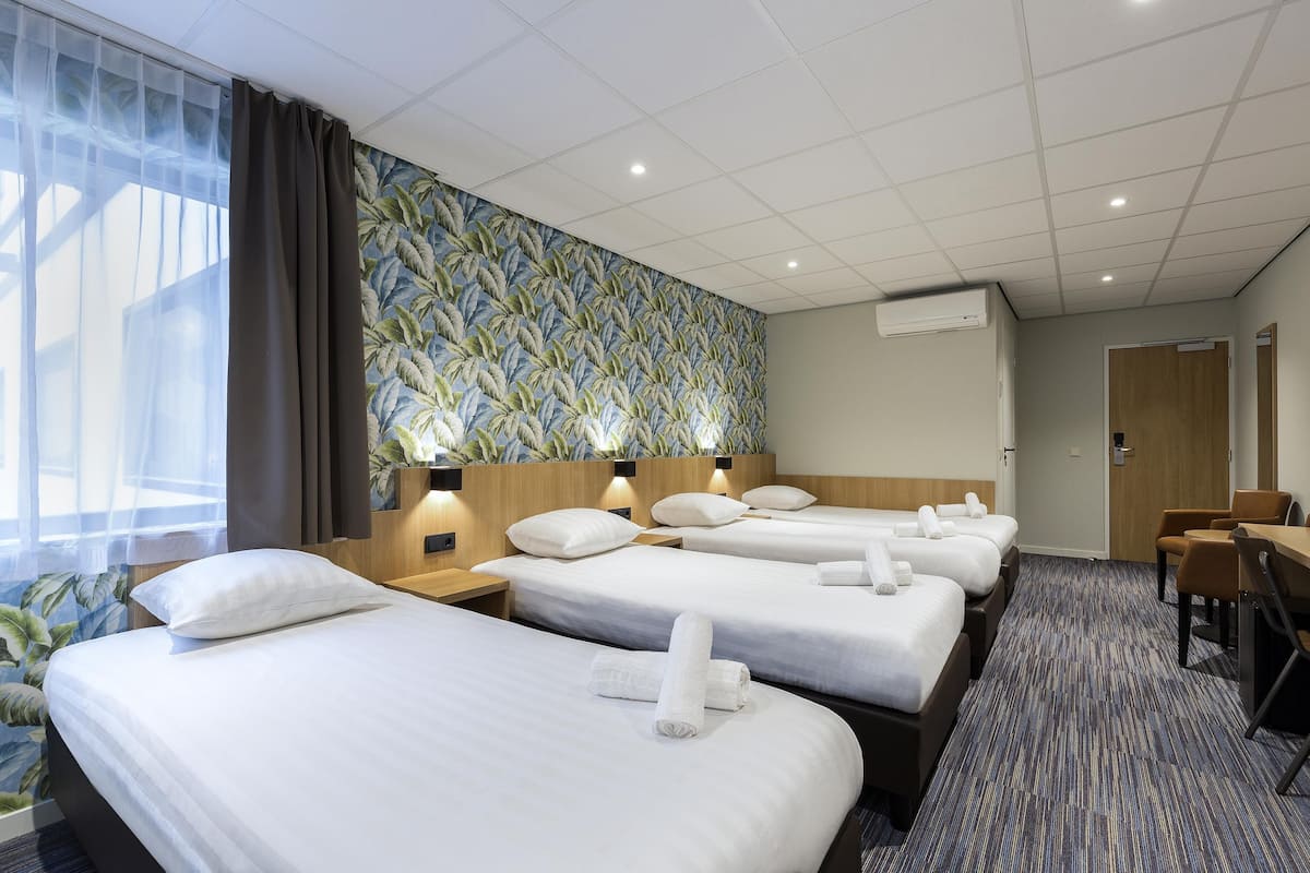 A hotel room with four beds, a large window with curtains, a wall with a leafy pattern, and a carpeted floor.