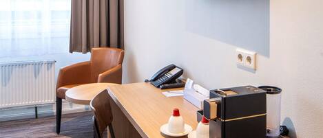 A hotel room with a wooden desk, two chairs, a flat-screen TV mounted on the wall, a telephone, a coffee machine, and a radiator.