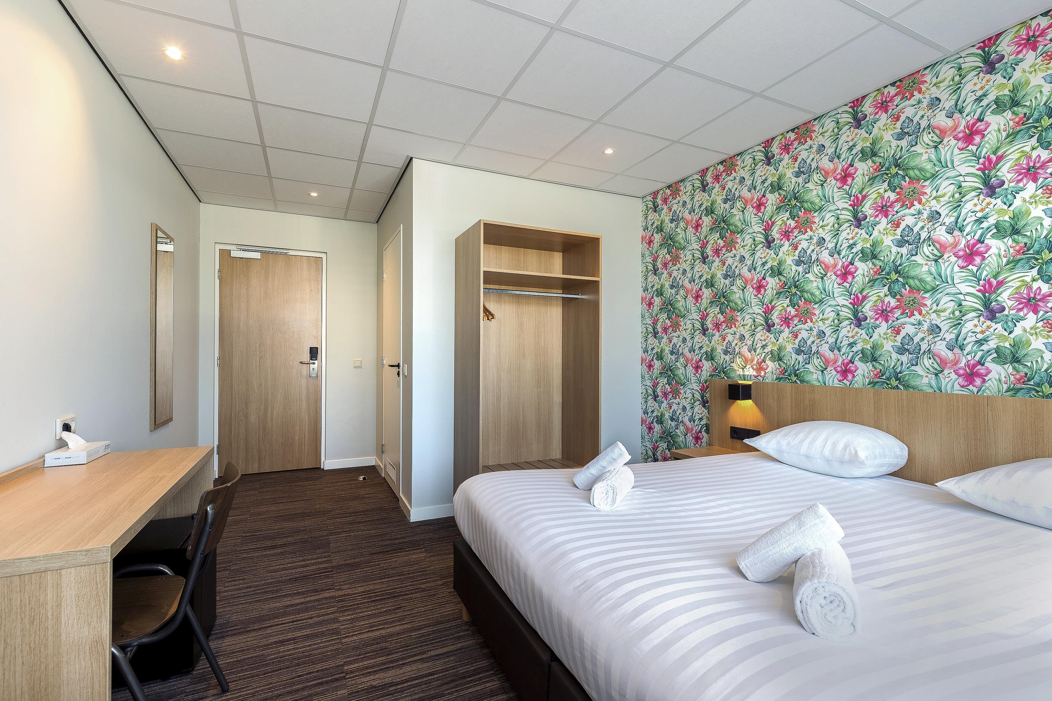 A hotel room with a bed, a desk, a chair, a wardrobe, and a floral wallpaper.
