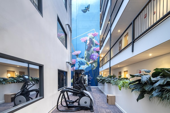 Un pasillo moderno de hotel con equipo de ejercicio, ventanas amplias y plantas de interior.