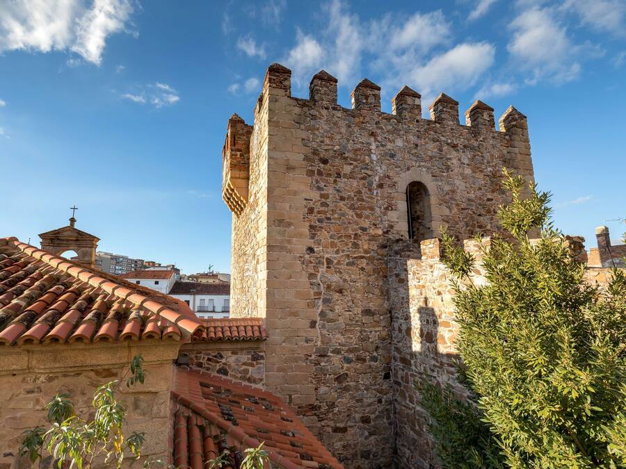 Una torre de piedra histórica con una cruz en la parte superior, rodeada de edificios con tejados de tejas.