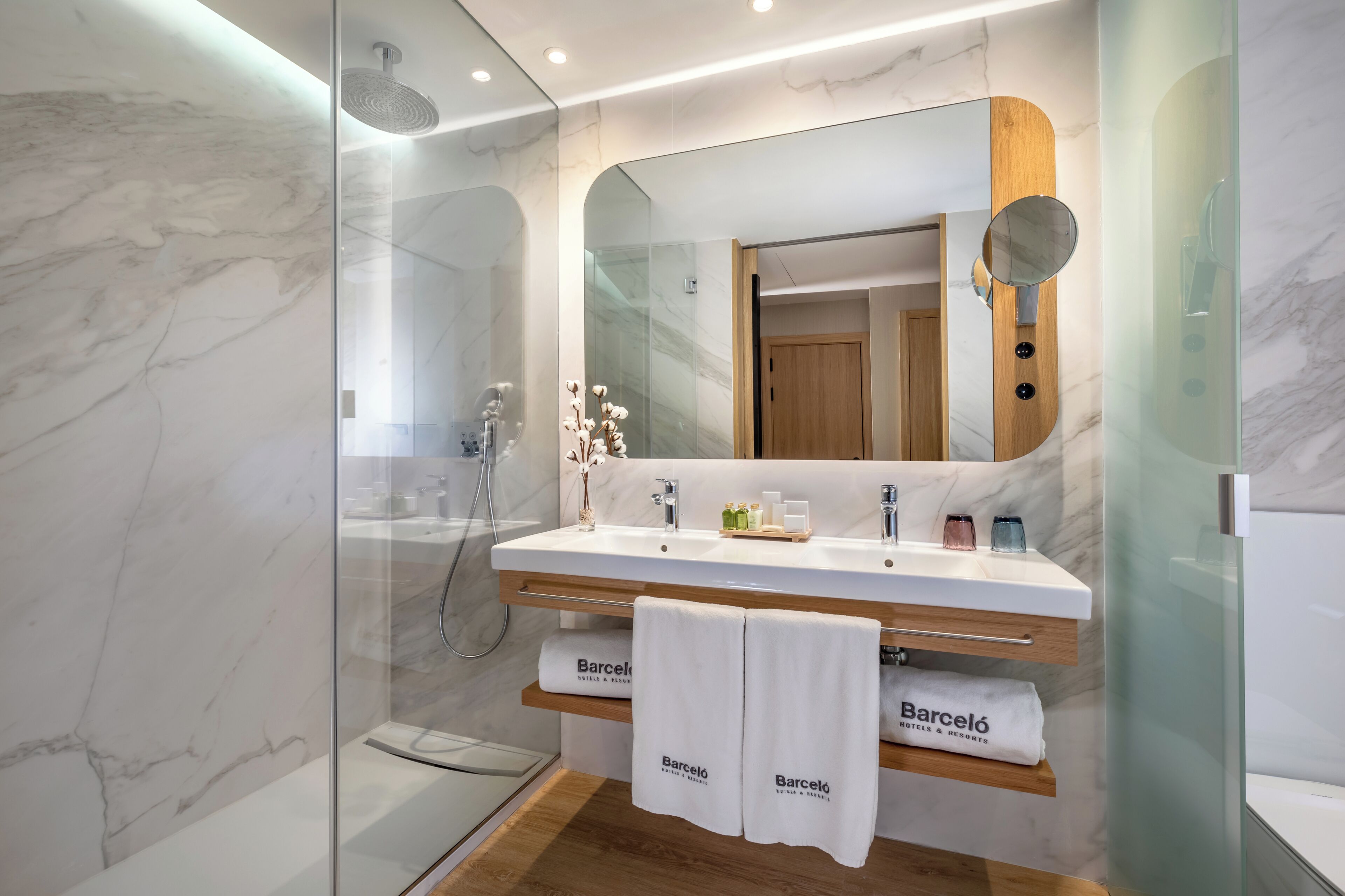 Un baño moderno con ducha envuelta en vidrio, pared con mosaicos de mármol, tocador de madera con espejo y dos toallas con el logotipo de 'Barceló Hotels & Resorts'.
