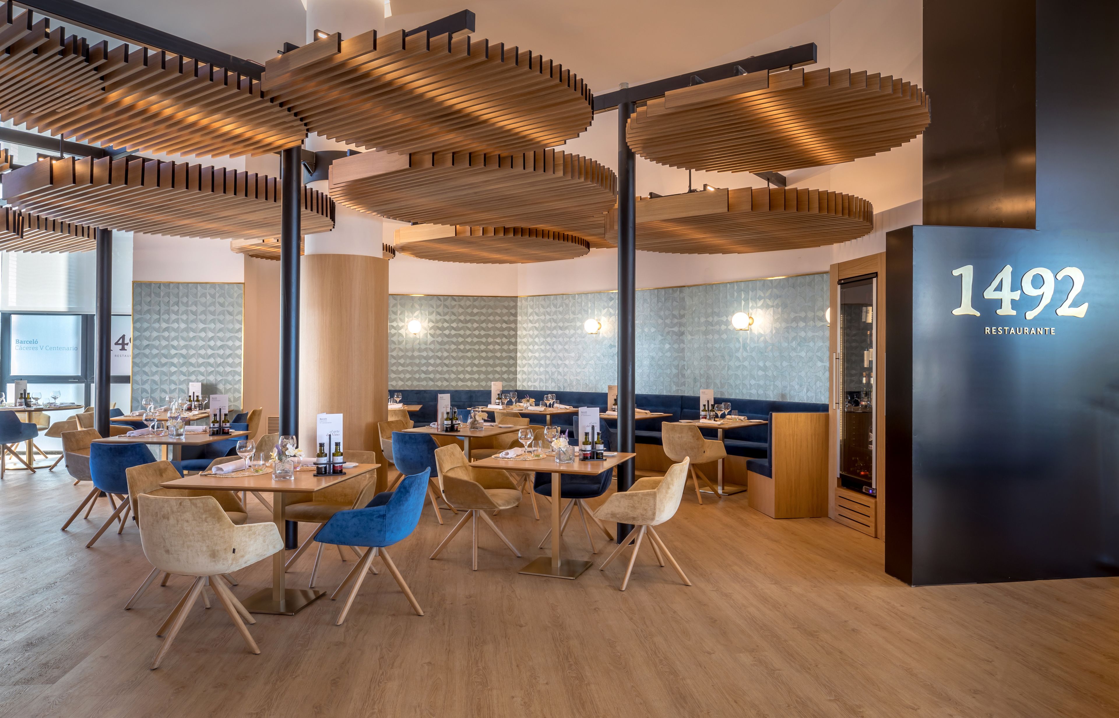 A modern restaurant interior with wooden ceiling structures, blue and beige chairs, and a black wall with the number 1492.