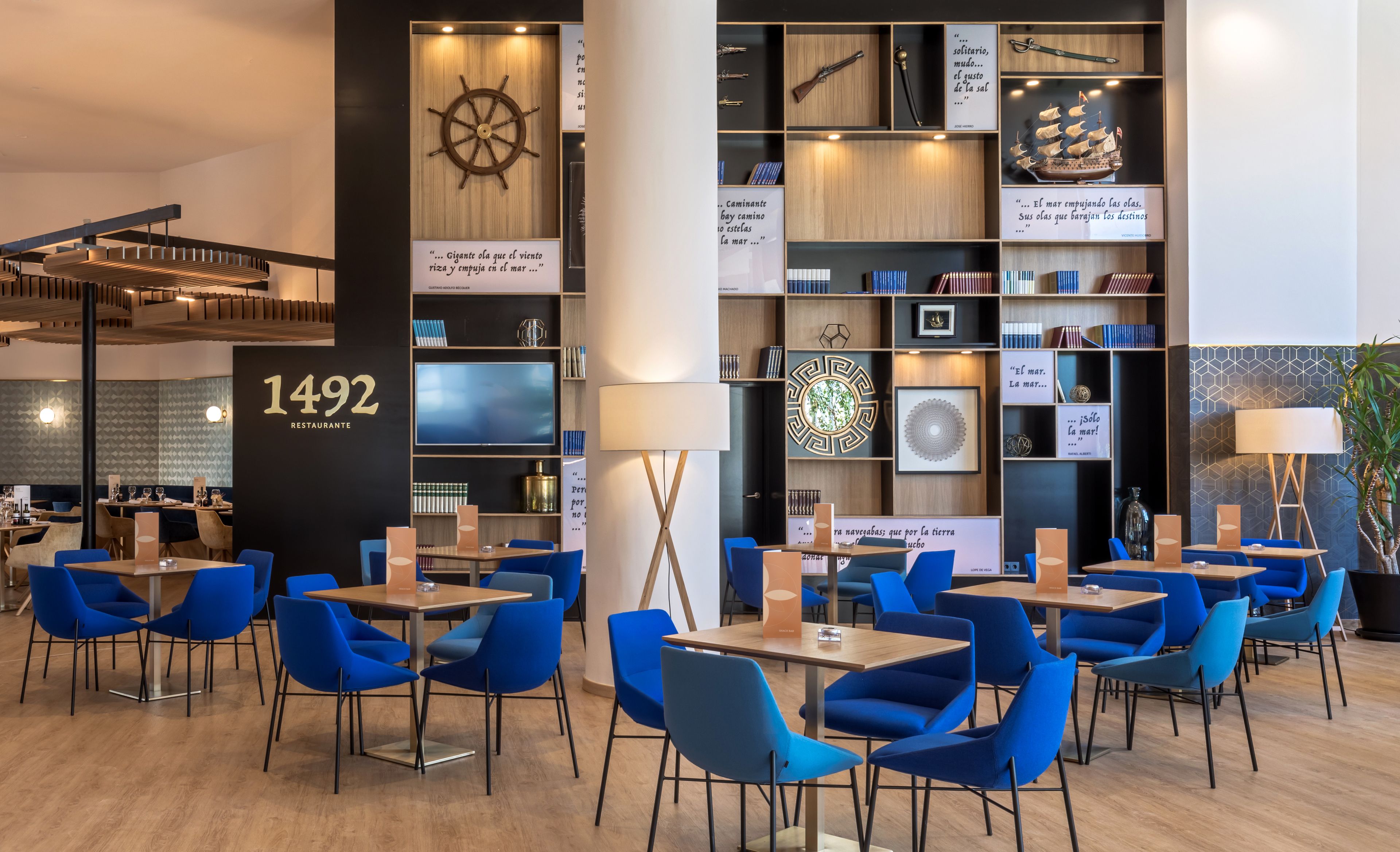 Un interior de restaurante moderno con sillas azules, mesas de madera y una gran pared que muestra varias citas enmarcadas y una rueda de barco.