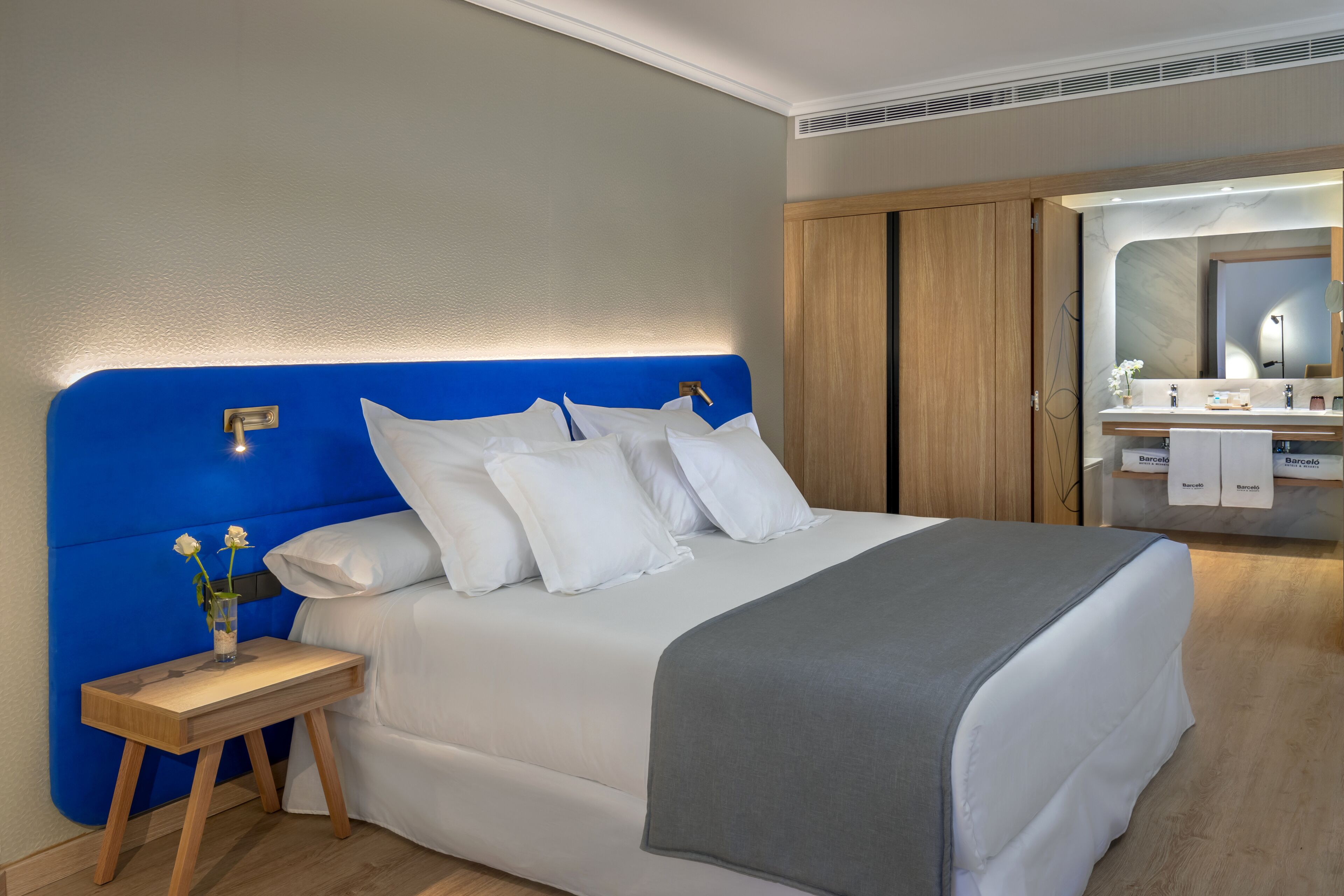 Una habitación de hotel moderna con una cama grande, una mesita de noche con un florero, y un baño visible a través de una puerta abierta.
