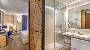 A modern hotel room with a wooden partition, a glass shower enclosure, a white bed, a desk with a chair, and a large mirror.