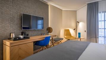 Una habitación de hotel moderna con una cama grande, un escritorio de madera con un televisor de pantalla plana, una silla azul, una mesita con una lámpara y vistas a la ciudad a través de amplias ventanas.