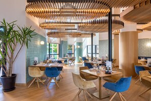 A modern dining area with wooden ceiling panels, potted plants, and a variety of seating arrangements. - Barceló Cáceres V Centenario (Cáceres)