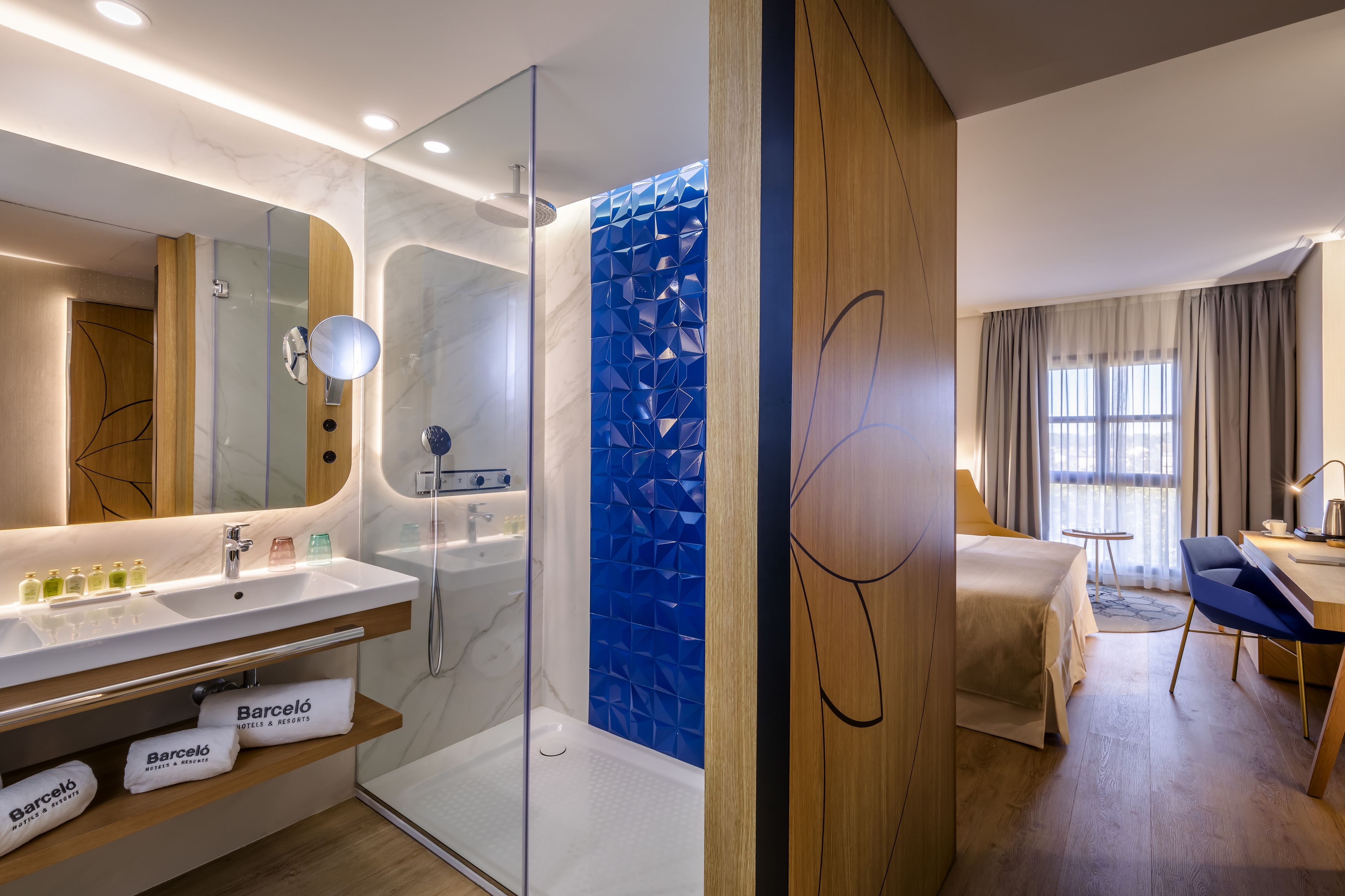 A modern hotel room with a glass-enclosed shower, a large mirror, a wooden vanity with a sink, and a bed with a view of the city.