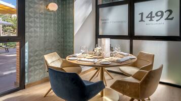 A dining area with a round table set for four, surrounded by four chairs, and a large window with the number 1492 visible.