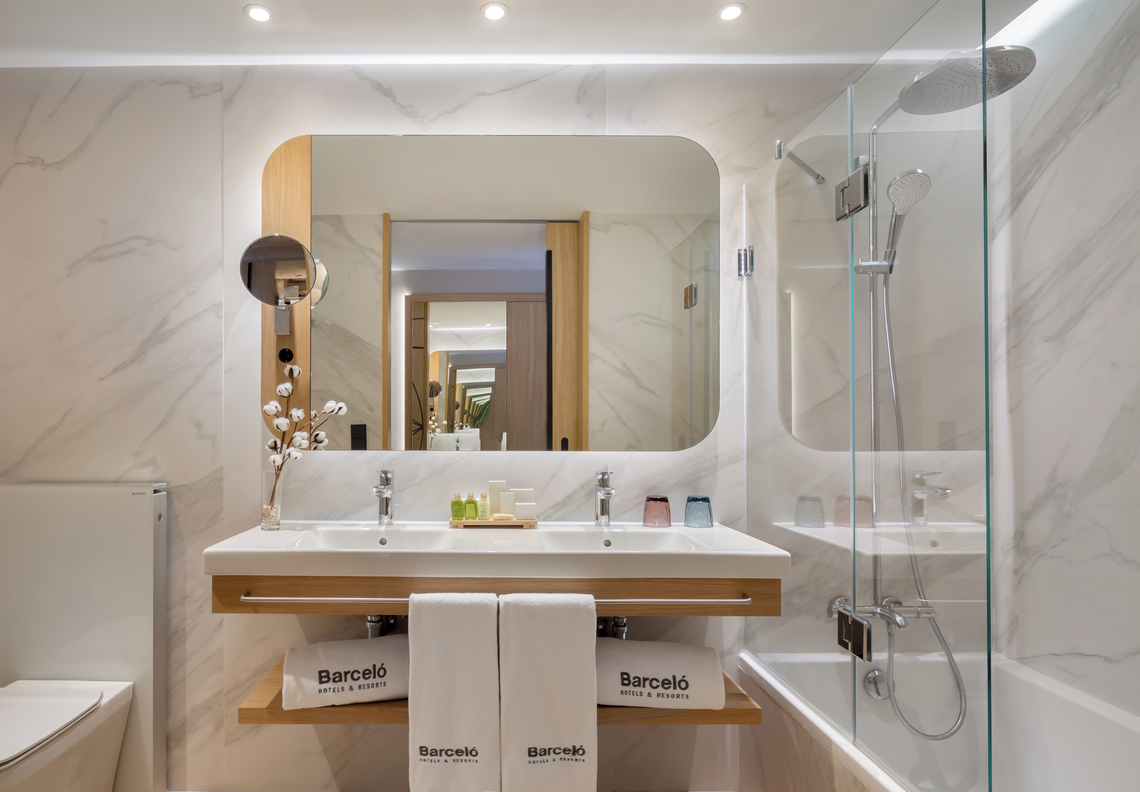 Uma casa de banho moderna com uma pia dupla, um espelho grande, cabine de chuveiro com portas de vidro e toalhas da marca 'Barceló Hotels & Resorts'.