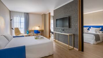 Una habitación de hotel moderna con una cama grande, un escritorio con silla, un televisor montado en la pared y una zona de descanso separada con cama.