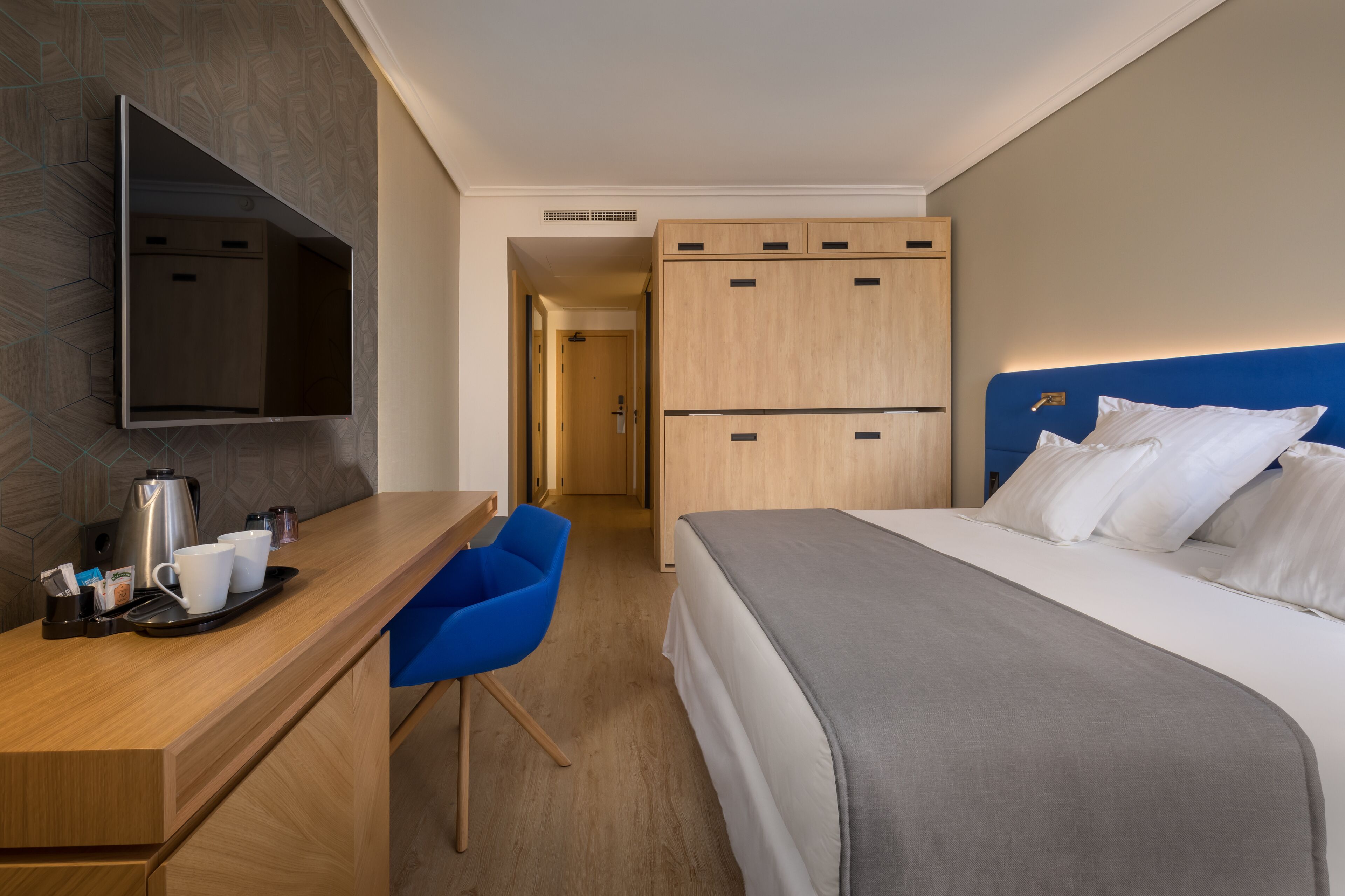 Una habitación de hotel con una cama, un escritorio con cafetera, una silla y un televisor montado en la pared.