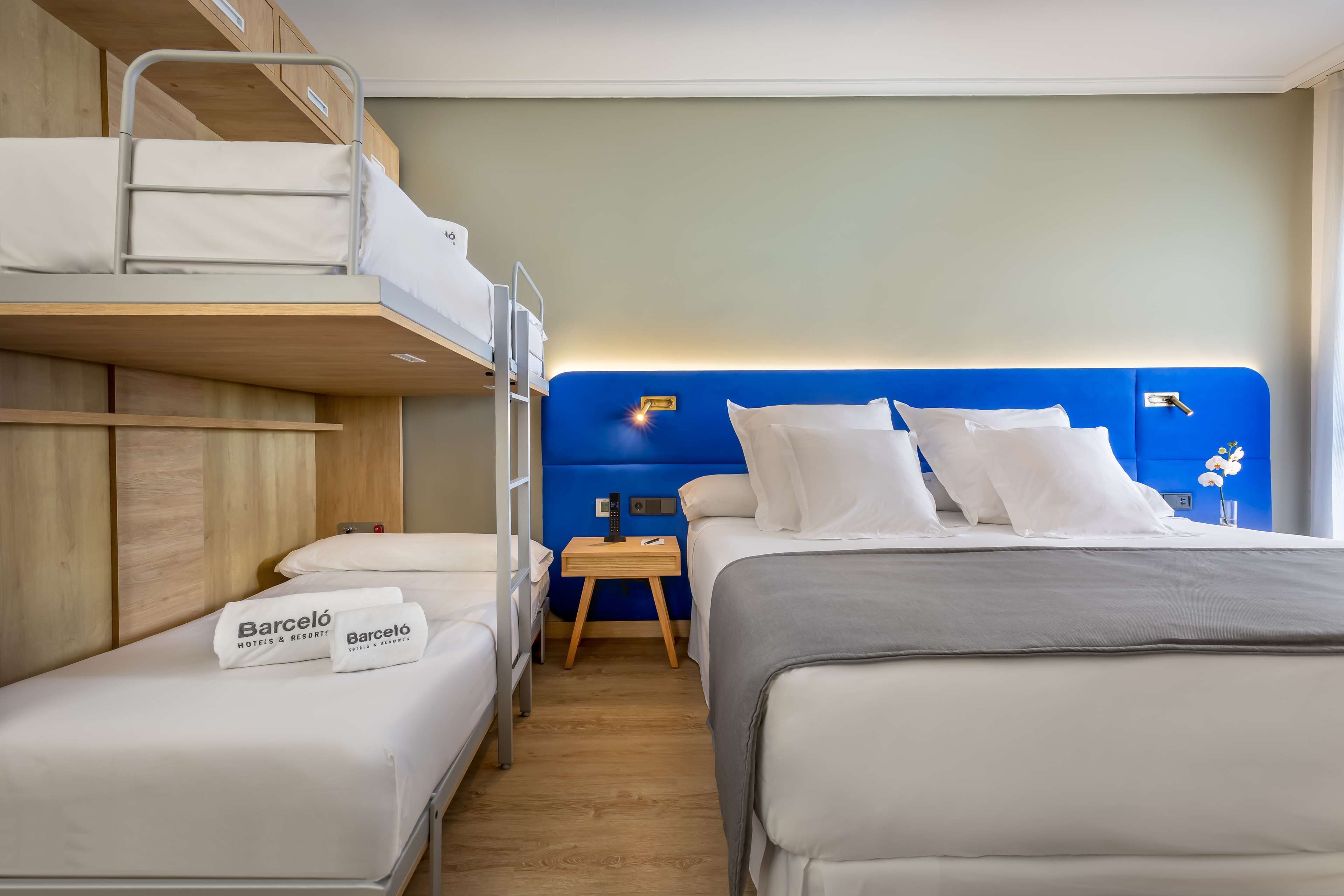 A hotel room with a bunk bed, a single bed, a nightstand with a lamp, and a small table.