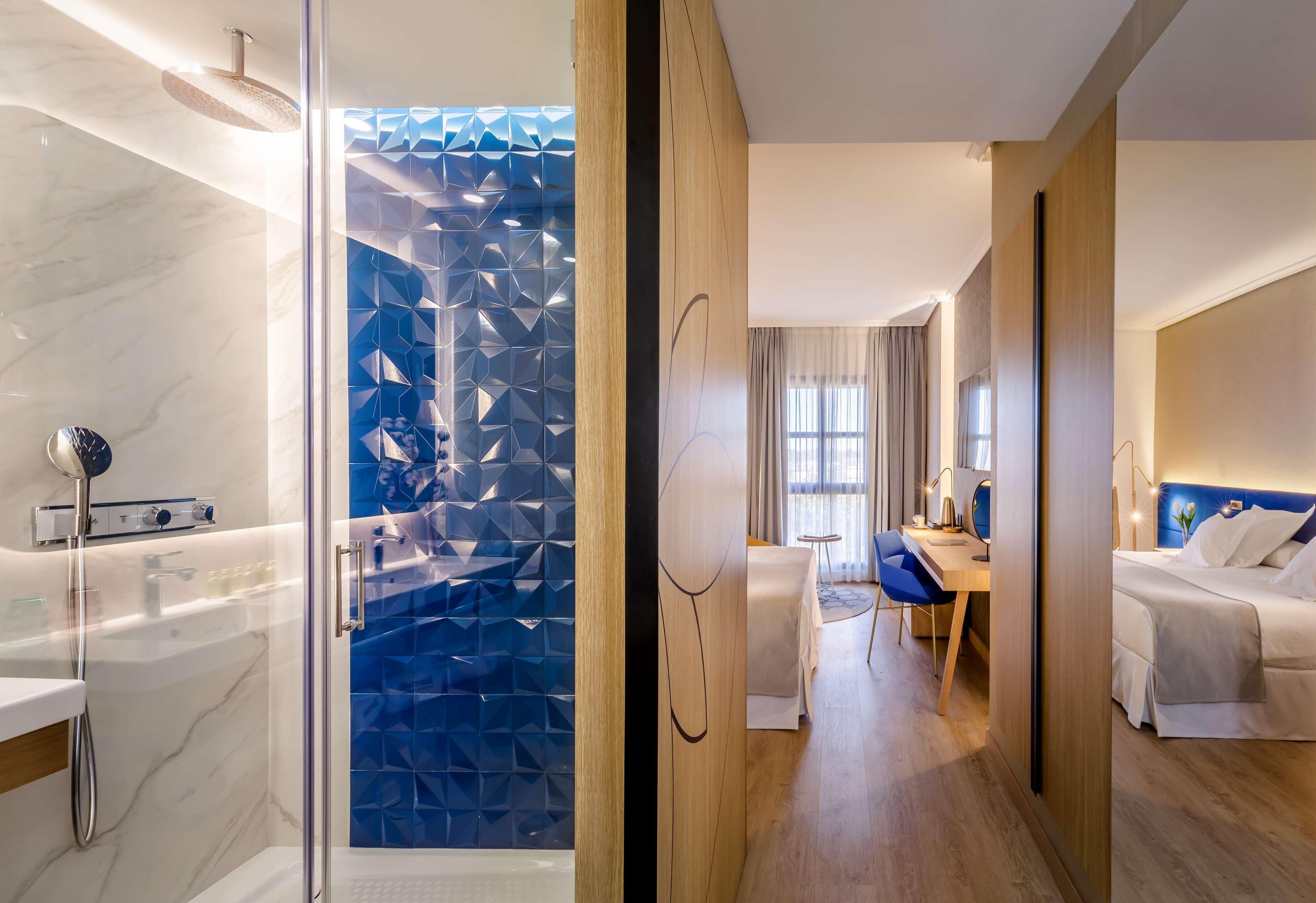 A modern hotel room with a glass shower enclosure, a wooden desk, a bed, and a view through a geometric-patterned glass door.