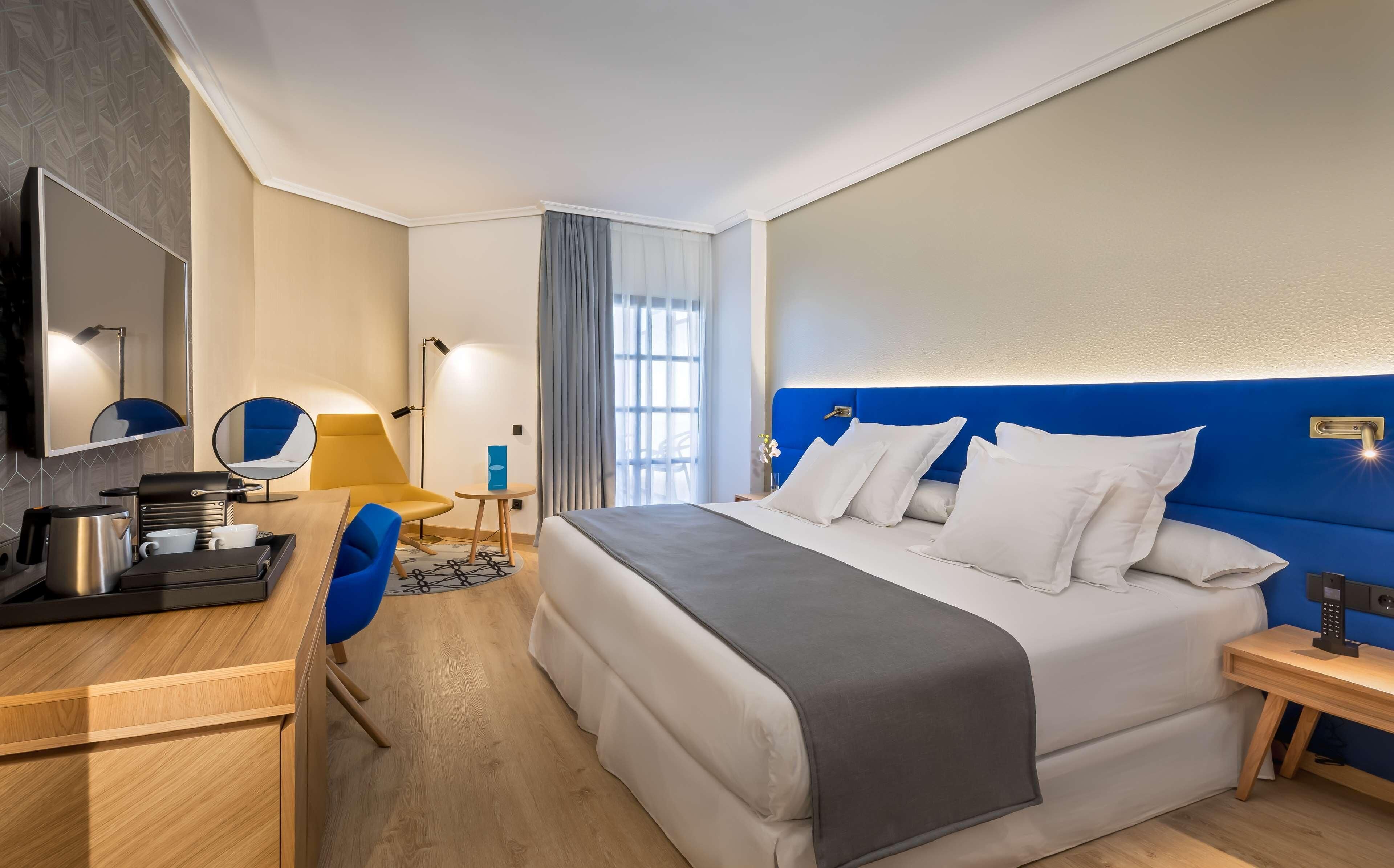 A hotel room with a bed, bedside table, desk, chair, lamp, and a view of the outside.