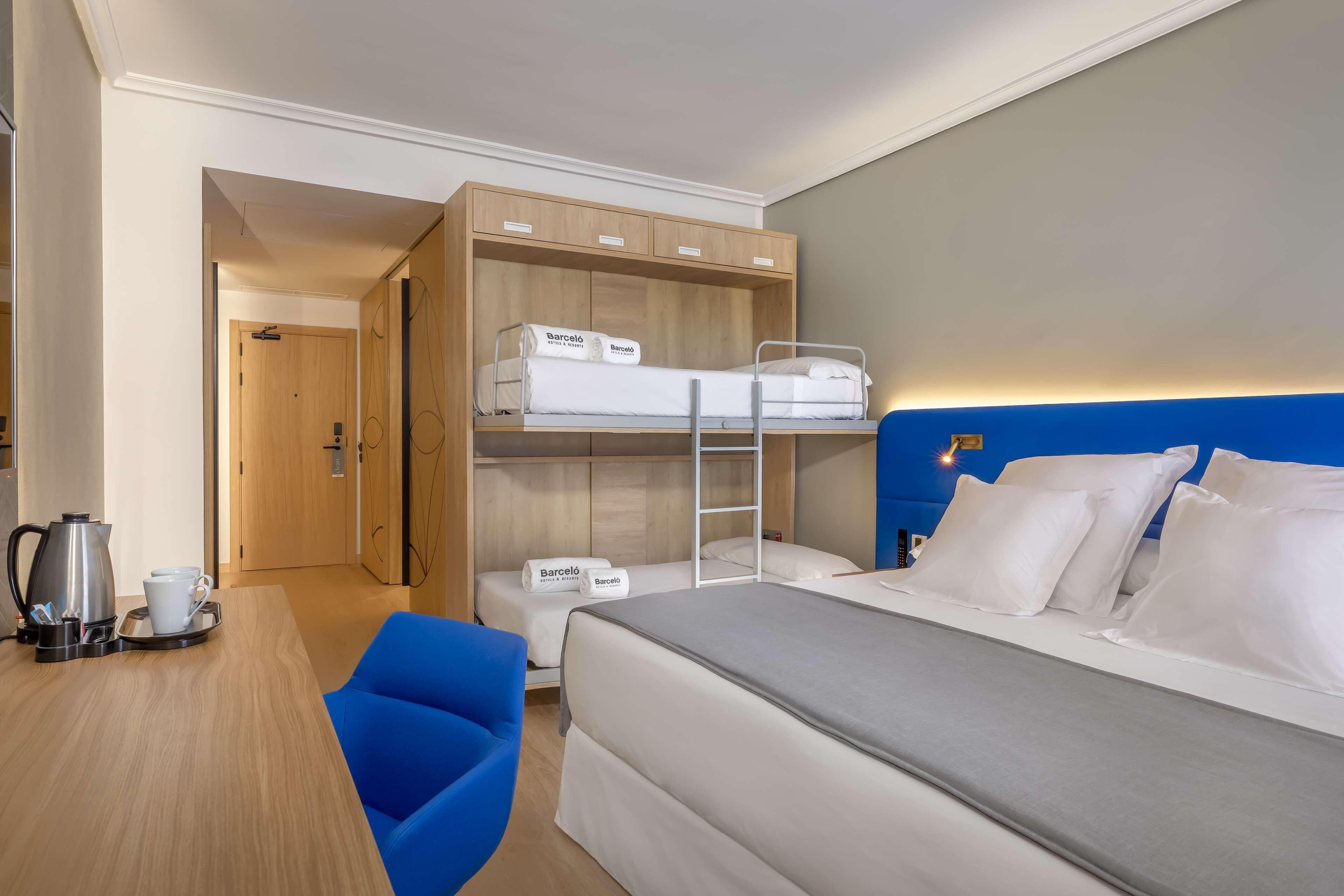 A hotel room with a bunk bed, a single bed, a desk with a coffee maker, and a blue chair.