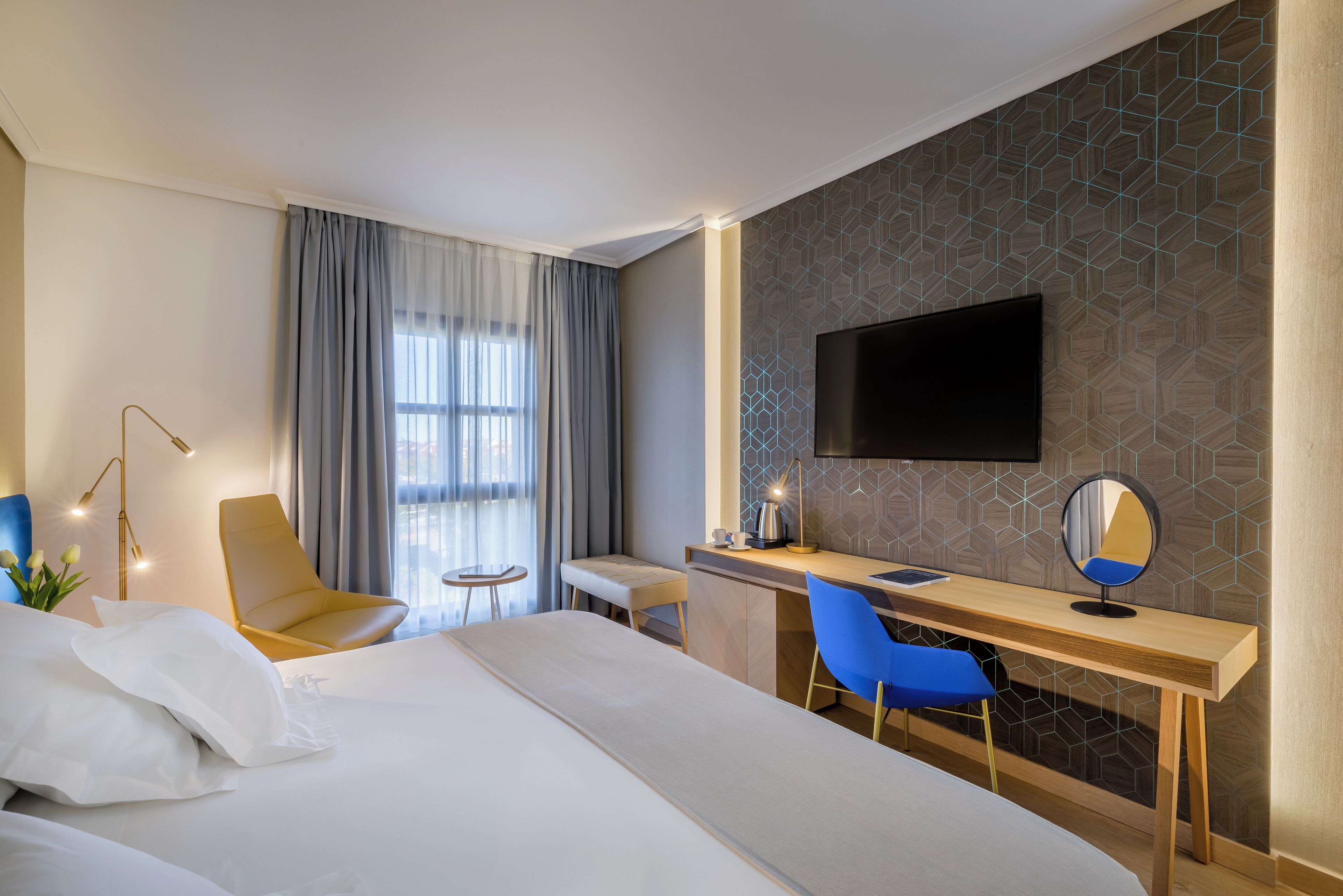 A hotel room with a bed, a desk, a chair, a TV, and a window with curtains.
