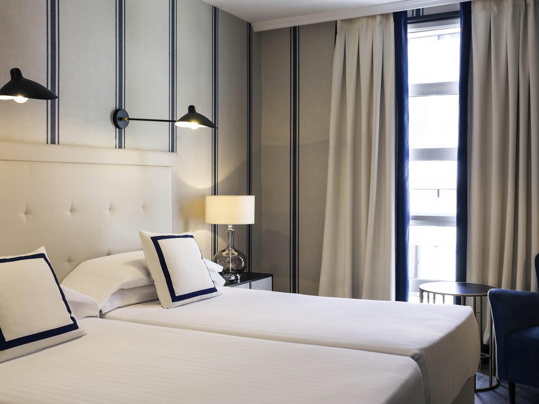 A hotel room with two beds, a nightstand with a lamp, a small table, and a view of the outside through a window with curtains.