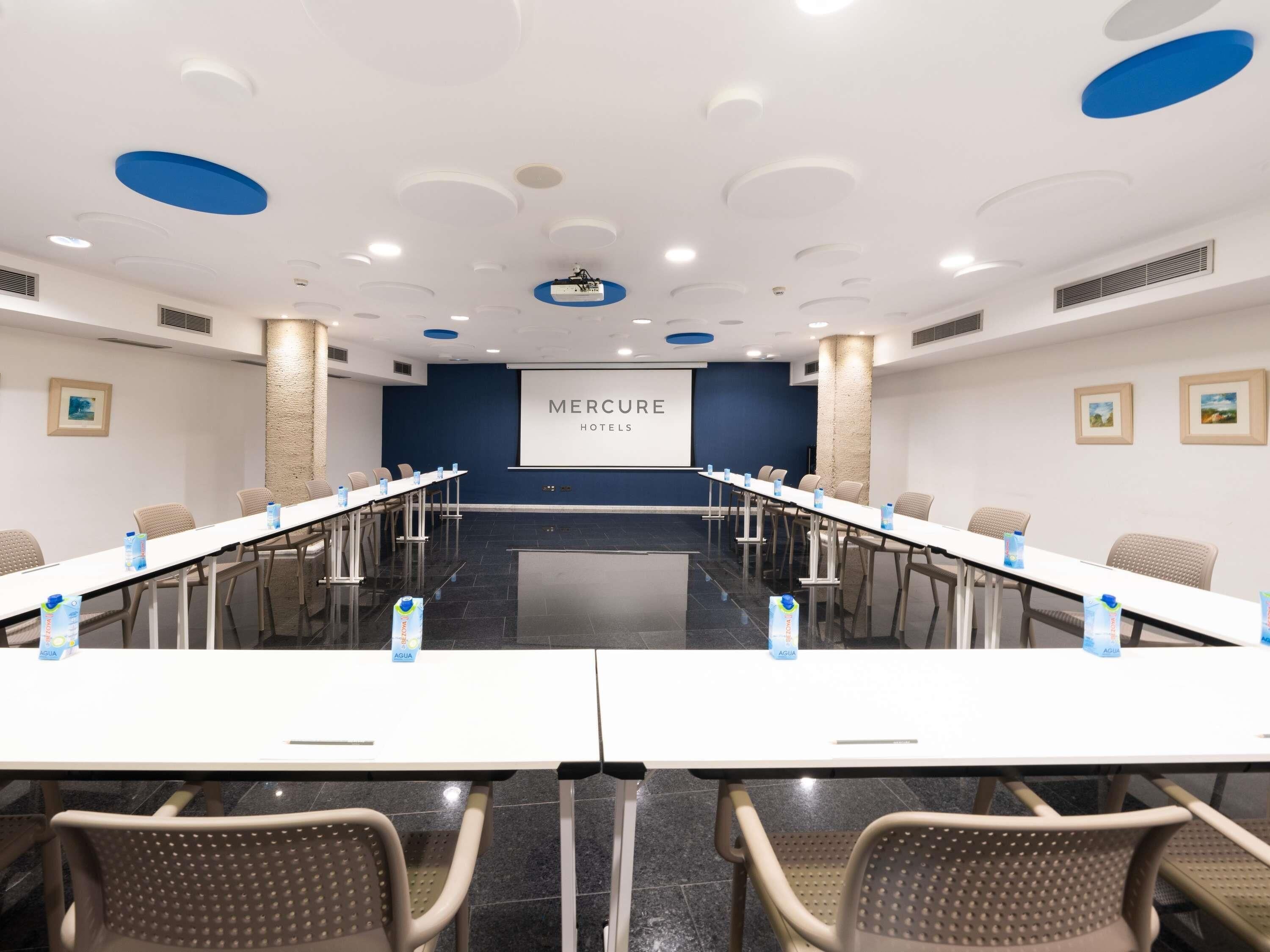 A conference room with a long table, chairs, and a screen displaying 'MERCURE HOTELS'.