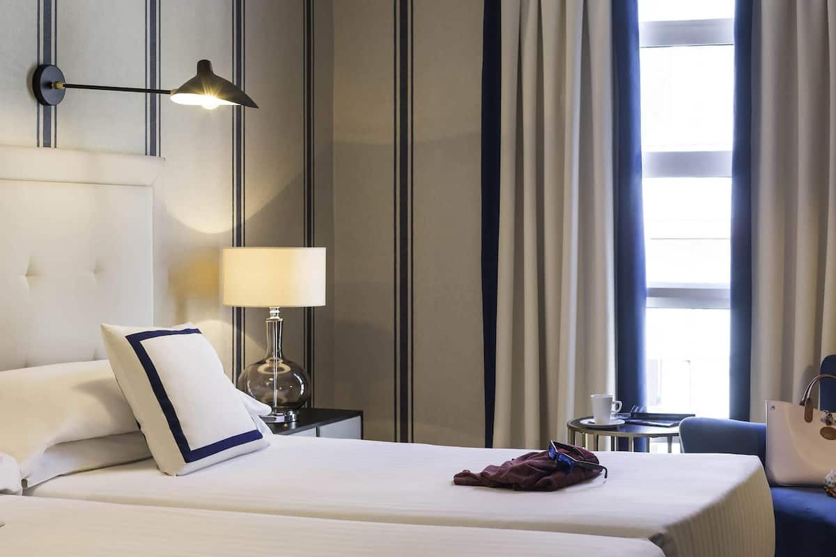 A hotel room with two beds, a nightstand with a lamp, a chair, and a view of the outdoors through a window.
