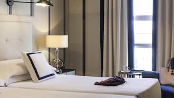 A hotel room with two beds, a nightstand with a lamp, a chair, and a view of the outdoors through a window.